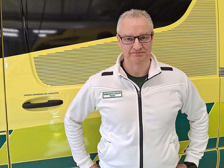 lääkäri seisoo ambulanssin edessä ja katsoo suoraan kameraan