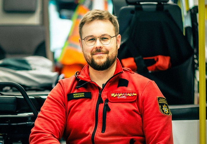 ensihoitaja istuu ambulanssin edessä ja hymyilee suoraan kameralle