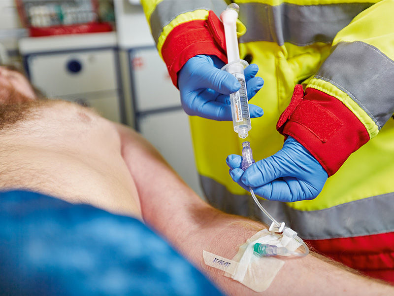 Potilas, joka on peitetty sinisellä peitteellä vatsan keskeltä, makaa ambulanssissa. Potilaan vasenta kyynärvartta kanyloidaan vihreällä Introcan Safety® 3 -kanyylilla. Caresite-venttiili ja infuusioletku sekä valkoinen liukupuristin on liitetty. Kiinnitys haavateipillä. Ensihoidon työasussa ja sinisissä käsineissä oleva henkilö pitää täytettyä ruiskua oikeassa kädessään. Vasemmalla kädellä pidetään letkua venttiilin alla.